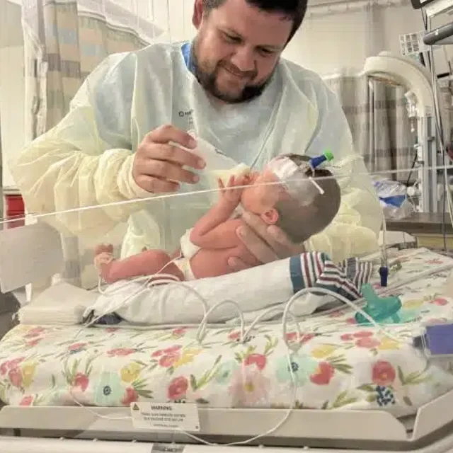 A father in a hospital gown feeding his premature baby in a NICU incubator