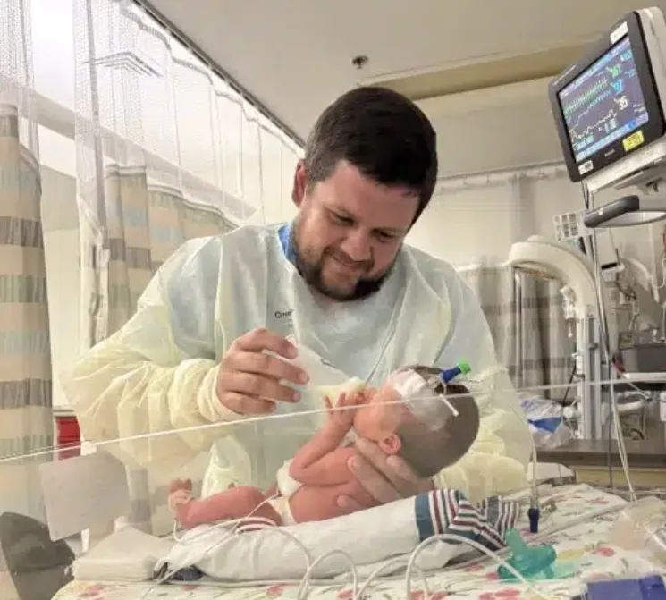 A father in a hospital gown feeding his premature baby in a NICU incubator